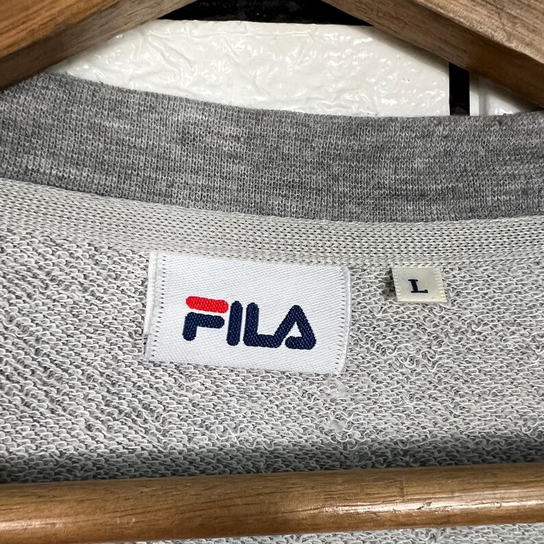 May include: Close-up of a gray FILA sweatshirt. The white label with the FILA logo in red and navy blue is visible. The size tag shows a size "L". The sweatshirt is on a wooden hanger.
