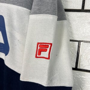 May include: A color-blocked sweatshirt featuring a navy blue, white, and gray design. The white section has a red embroidered logo. The sweatshirt has a crew neck and long sleeves. The logo is a red square with a white "F" and "L" inside.