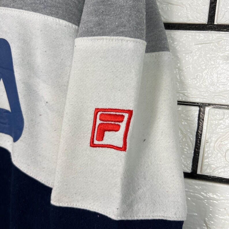 May include: A color-blocked sweatshirt featuring a navy blue, white, and gray design. The white section has a red embroidered logo. The sweatshirt has a crew neck and long sleeves. The logo is a red square with a white "F" and "L" inside.