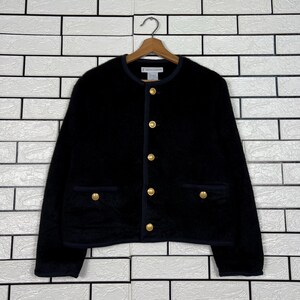May include: A black jacket with gold buttons and navy blue trim. The jacket has two front pockets and a round neckline. The jacket is hanging on a wooden hanger against a white brick wall.