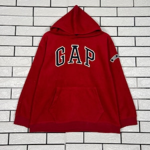 Pode incluir: Um moletom vermelho com capuz com a palavra "GAP" em letras brancas e azul marinho no peito. O moletom tem um bolso frontal e um pequeno patch na manga que diz "EST."