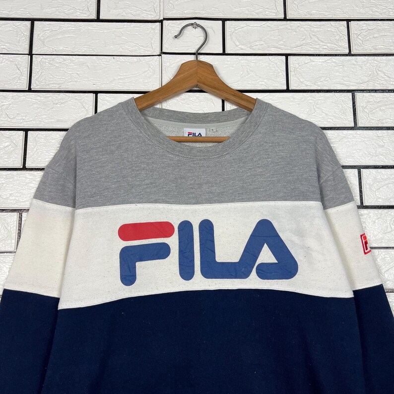 May include: A color-blocked sweatshirt in gray, white, and navy blue, featuring the brand name "FILA" in large, bold letters across the chest. The logo is in red and blue. The sweatshirt is hanging on a wooden hanger.