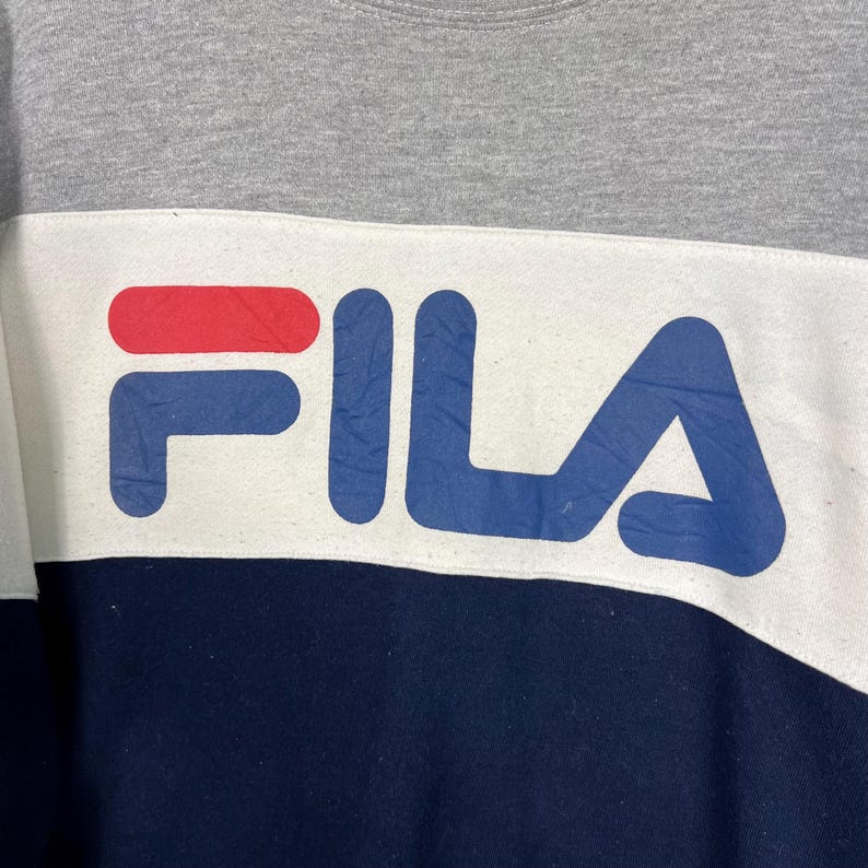 May include: A gray, white, and navy blue sweatshirt with the brand name "FILA" in large, bold letters across the chest. The logo is in red and blue. The sweatshirt has a color block design.