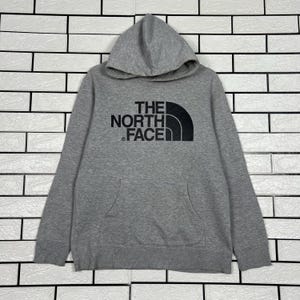 May include: A gray hooded sweatshirt with a kangaroo pocket and the text "THE NORTH FACE" in black. The sweatshirt has a drawstring hood and long sleeves. The North Face logo is a black half-circle over three vertical lines.