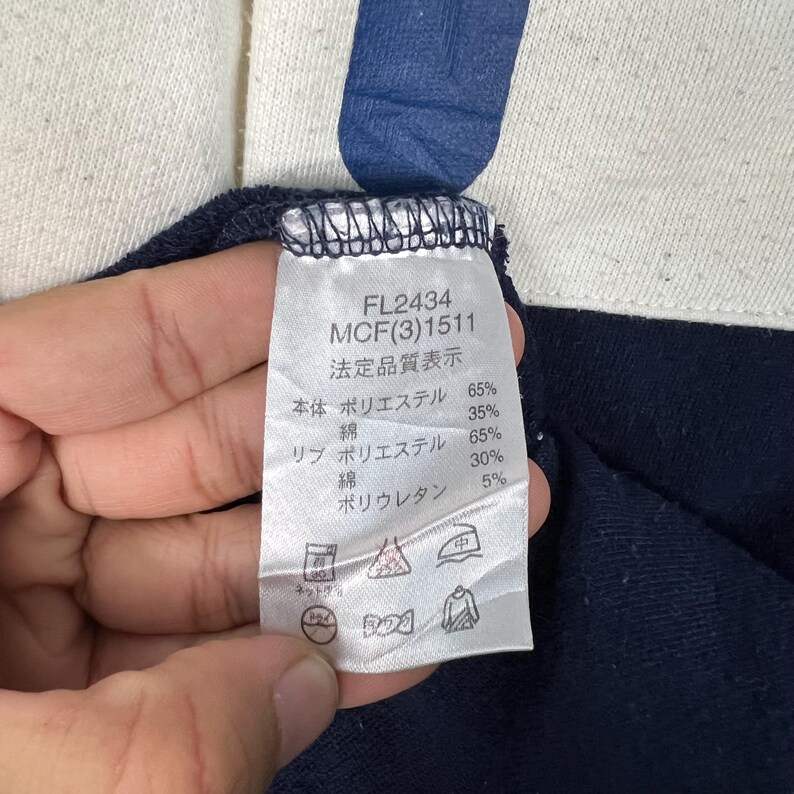 May include: Close-up of a clothing tag with the text "FL2434 MCF(3)1511" and Japanese characters. The tag lists fabric composition percentages: 65% polyester, 35% cotton, 65% polyester, 30% cotton, and 5% polyurethane. The tag is attached to a navy blue garment.