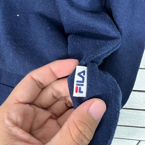 May include: Close-up of a navy blue sweatshirt with a white tag featuring the brand name "FILA" in a stylized font. The tag has red accents. The fabric appears soft and slightly textured.