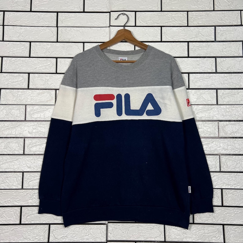 May include: A color-blocked sweatshirt with the brand name "FILA" in large, navy blue letters across the chest. The top portion is gray, with a white band across the chest, and the bottom is navy blue. A small red "F" logo is on the right sleeve.