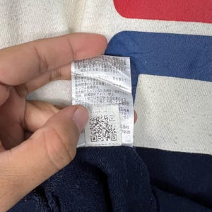 May include: Close-up of a tag with Japanese text, a QR code, and the number 12-20. The tag is attached to a navy blue and white fabric with a red stripe. The fabric appears to be part of a garment.