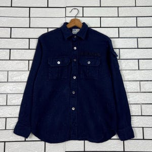 Puede incluir: Una camisa azul marino de manga larga con dos bolsillos en el pecho y cierre de botones. La camisa tiene cuello y un pequeño parche en el pecho izquierdo. La camisa está colgada en una percha de madera contra una pared de ladrillos blancos.