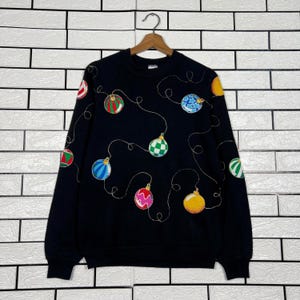 May include: Black sweatshirt with embroidered Christmas ornaments in various colors, including red, green, blue, and yellow. The ornaments are connected by gold thread-like lines, creating a festive design. The sweatshirt is hanging on a wooden hanger.