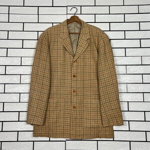 May include: A tan and brown plaid blazer with three buttons and two front pockets. The jacket is hanging on a wooden hanger against a white brick wall. The blazer has a classic, tailored design.