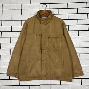 Peut inclure: Veste de couleur beige avec un col montant et plusieurs poches. La veste a une fermeture éclair et un petit logo près du col. Elle est suspendue à un cintre en bois contre un mur de briques blanches.