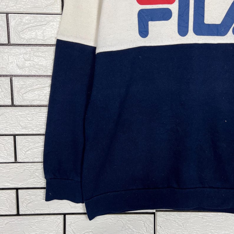 May include: A navy blue and white sweatshirt with the word "FILA" in large, bold letters across the chest. The sweatshirt has a color-block design, with a white upper section and a navy blue lower section. The sleeves are navy blue.