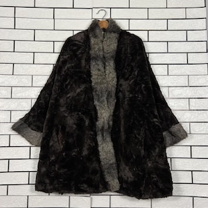 May include: A dark brown faux fur coat with a gray fur collar and trim, hanging on a wooden hanger. The coat has a wide, flowing design and is displayed against a white brick wall.