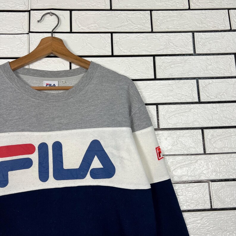 May include: A gray, white, and navy blue FILA sweatshirt. The sweatshirt has a color block design with the FILA logo in large letters across the chest. The long-sleeved shirt is hanging on a wooden hanger.