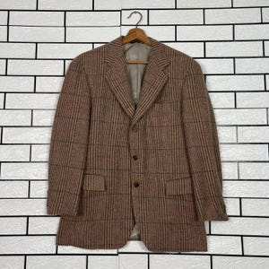 May include: A brown plaid blazer with a notched lapel, three-button closure, and two front pockets. The jacket is hanging on a wooden hanger against a white brick wall.