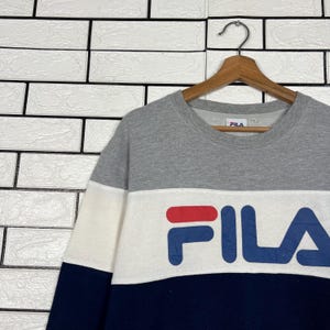 May include: A gray, white, and navy blue Fila sweatshirt. The sweatshirt has a color block design with the Fila logo in navy blue and red. The sweatshirt is hanging on a wooden hanger against a white brick wall.