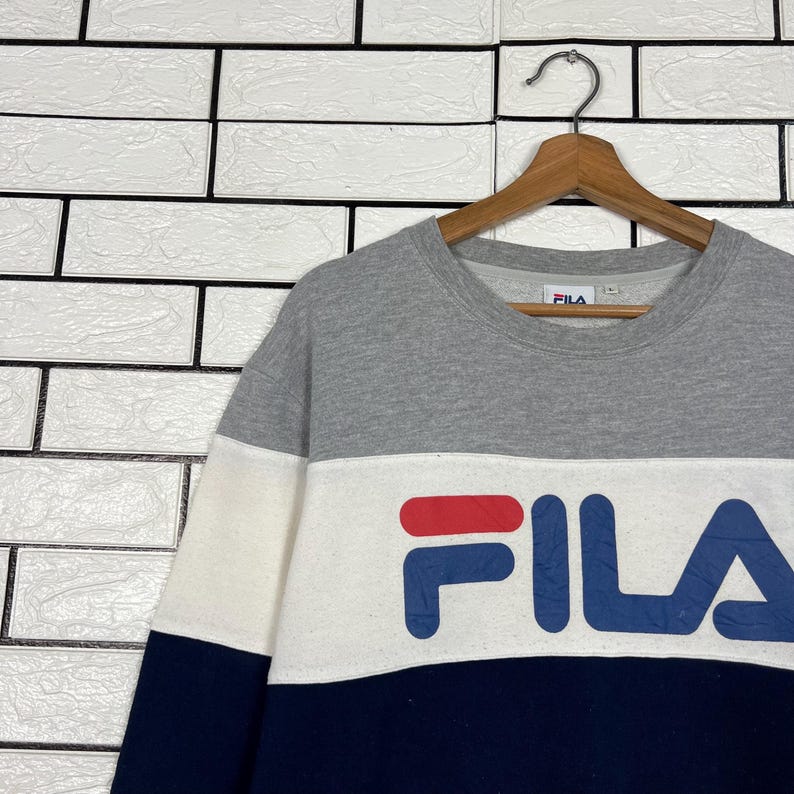 May include: A gray, white, and navy blue Fila sweatshirt. The sweatshirt has a color block design with the Fila logo in navy blue and red. The sweatshirt is hanging on a wooden hanger against a white brick wall.