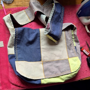 Peut inclure: Sac à bandoulière patchwork dans les tons de bleu, beige et gris, avec une longue bandoulière. Le sac est doté d'une fermeture à rabat et est cousu avec du fil jaune. Le sac est en tissu.