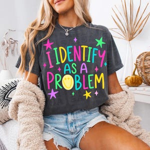 May include: A charcoal gray short-sleeve t-shirt featuring the text I IDENTIFY AS A PROBLEM in bright yellow, pink, and teal lettering. The design includes a yellow smiley face icon and several small star shapes in pink, yellow, and green.