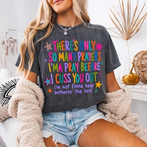 May include: A charcoal gray short-sleeve t-shirt featuring colorful text that reads There's Only So Many Prayers I'ma Pray Before I Cuss You Out and i'm not finna keep botherin' the lord, accented with small star and leopard print graphics.