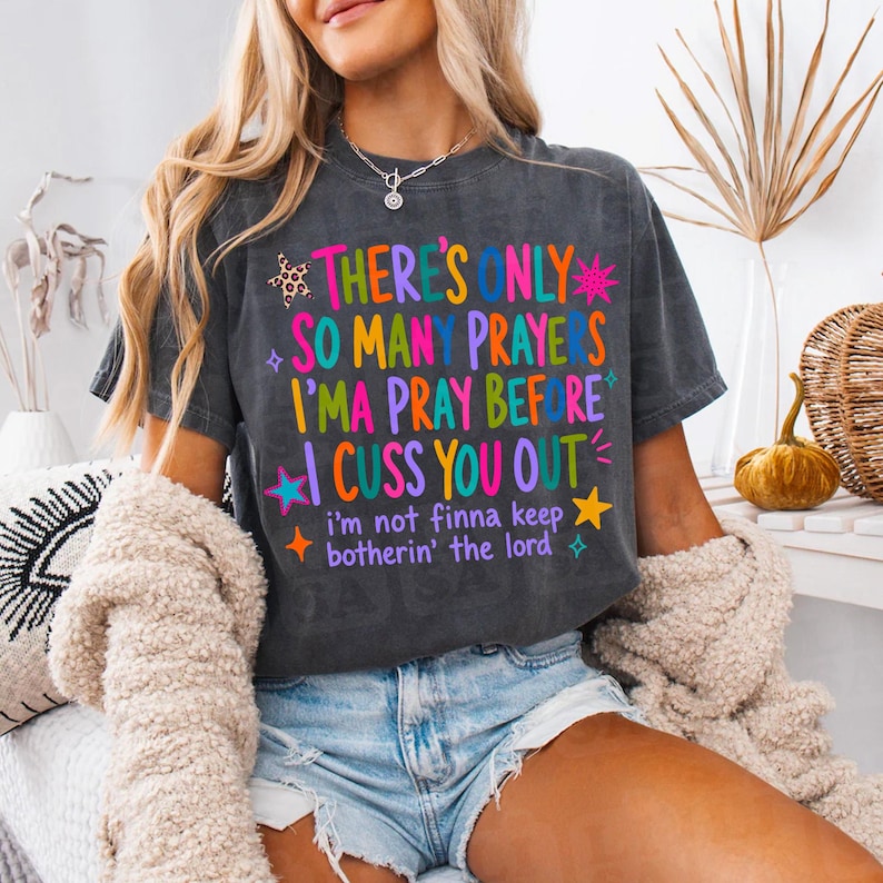 May include: A charcoal gray short-sleeve t-shirt featuring colorful text that reads There's Only So Many Prayers I'ma Pray Before I Cuss You Out and i'm not finna keep botherin' the lord, accented with small star and leopard print graphics.