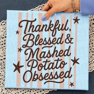 May include: A square canvas sign with a light blue background and vertical light pink stripes. The text in dark brown script reads Thankful, Blessed & Mashed Potato Obsessed, accented by several dark brown stars scattered around the lettering.