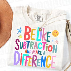 May include: Cream-colored t-shirt with the text "BE LIKE SUBTRACTION AND MAKE A DIFFERENCE" in various bright colors. The shirt also features a gold necklace and a brown hat.