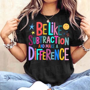 May include: Black t-shirt with the text "BE LIKE SUBTRACTION AND MAKE A DIFFERENCE" in colorful block letters. The shirt features a smiley face and star graphics. The shirt is a casual style.