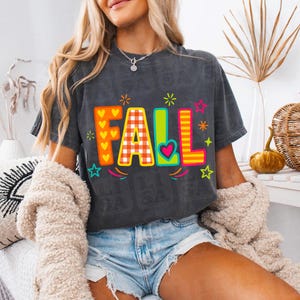 May include: A charcoal grey short-sleeve t-shirt featuring the word FALL in large, colourful, patterned block letters. The F has yellow hearts, the A has a red and white gingham check, the L has a heart, and the second L has orange stripes.