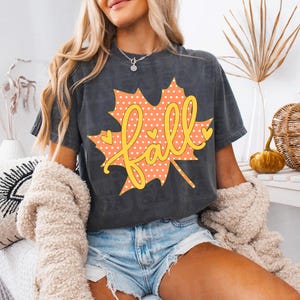 Może przedstawiać: Ciemnoszary t-shirt z krótkim rękawem z dużą grafiką pomarańczowego liścia klonu w białe kropki oraz żółtym napisem "fall" pisanym kursywą na środku. Wzór zawiera małe żółte serduszka zdobiące kształt liścia.