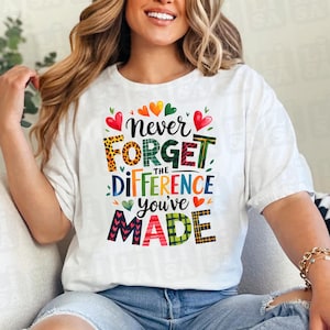 Può includere: T-shirt bianco con la scritta "Never Forget the Difference You've Made" in un carattere decorativo e colorato. Il design include cuori e vari motivi, creando un'atmosfera allegra e positiva. È un capo d'abbigliamento casual.