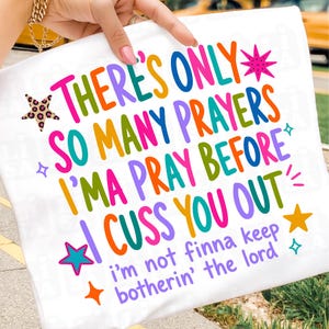 May include: A white t-shirt featuring colorful text that reads: THERE'S ONLY SO MANY PRAYERS I'MA PRAY BEFORE I CUSS YOU OUT and i'm not finna keep botherin' the lord. The design includes various multi-colored stars and small sparkle icons.