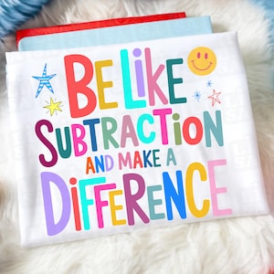 May include: White t-shirt with the text "BE LIKE SUBTRACTION AND MAKE A DIFFERENCE" in colorful letters. The design includes a smiley face, stars, and a starburst. The shirt is laying on a fluffy white surface.