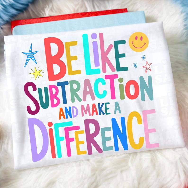 May include: White t-shirt with the text "BE LIKE SUBTRACTION AND MAKE A DIFFERENCE" in colorful letters. The design includes a smiley face, stars, and a starburst. The shirt is laying on a fluffy white surface.