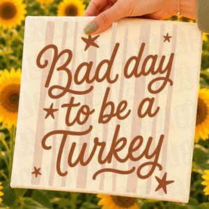 May include: A square canvas sign featuring a cream background with vertical light beige stripes. The text "Bad day to be a Turkey" is printed in a dark brown cursive font, accented by four small dark brown stars positioned around the text.