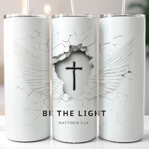 May include: White tumbler with a cracked design and angel wings. The center of the tumbler features a black cross and the text "BE THE LIGHT" and "MATTHEW 5:14". The tumbler has a straw.