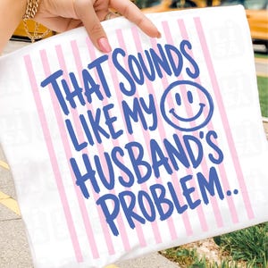 May include: White pillowcase with pink vertical stripes and the text "THAT SOUNDS LIKE MY HUSBAND'S PROBLEM.." in blue. A smiling face graphic is included in the text. The pillowcase is held by a person.