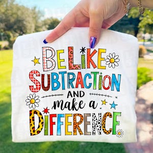 May include: White t-shirt featuring the phrase "BE LIKE SUBTRACTION AND make a DIFFERENCE" in a vibrant, multi-coloured design. The text is adorned with stars, daisies, and patterns. The shirt is held up by a hand.