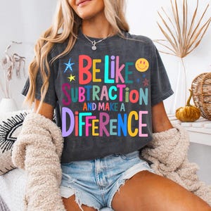 May include: Dark gray t-shirt with the text "BE LIKE SUBTRACTION AND MAKE A DIFFERENCE" in colorful letters. The shirt has a relaxed fit and is worn with denim shorts and a cream-colored cardigan. A necklace is visible.