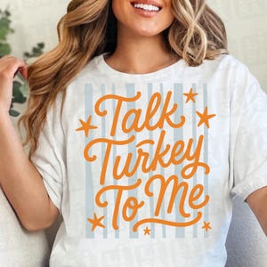May include: A white short-sleeved t-shirt featuring a graphic design with vertical light blue stripes. The text "Talk Turkey To Me" is printed in a bold orange cursive font, accented by several orange stars surrounding the lettering.