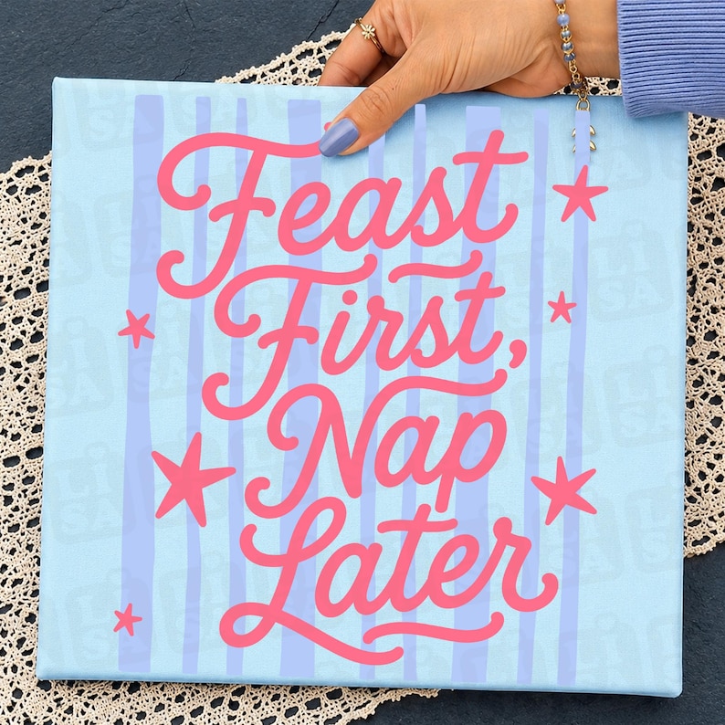 May include: A square light blue canvas sign featuring vertical lavender stripes and the pink cursive text Feast First, Nap Later. The design is accented with small pink stars scattered around the lettering, creating a decorative wall art piece.