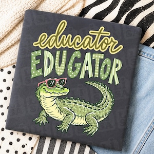May include: A dark grey t-shirt featuring a graphic of a green alligator wearing pink sunglasses. Above the alligator, the word educator is printed in yellow script, and the word EDUGATOR is printed below in large, green, patterned block letters.