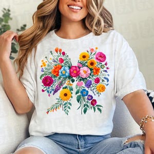 May include: White t-shirt featuring a vibrant floral bow design. The bow is composed of various colorful flowers, including pink, orange, yellow, and blue blooms, with green foliage. The shirt is a casual, comfortable style.