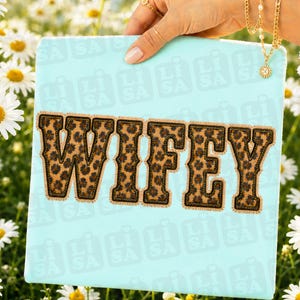 Könnte beinhalten: Eine hellblaue Leinwand mit dem Wort "WIFEY" in großen Buchstaben mit Leopardenmuster. Die Buchstaben haben einen braunen Rand und eine genähte Optik. Die Leinwand wird von einer Hand gehalten.