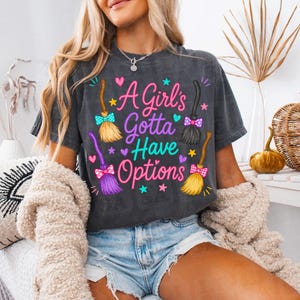May include: A charcoal gray short-sleeve t-shirt featuring the text A Girl's Gotta Have Options in pink and teal. The design includes four colorful witch broomsticks with patterned bows, surrounded by small pink and teal hearts and stars.