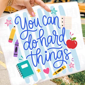 May include: A white t-shirt featuring the blue text You can do hard things, surrounded by colorful school-themed graphics including a yellow pencil, purple crayon, red apple, teal notebook, lined paper, and various pink, yellow, and teal stars and hearts.