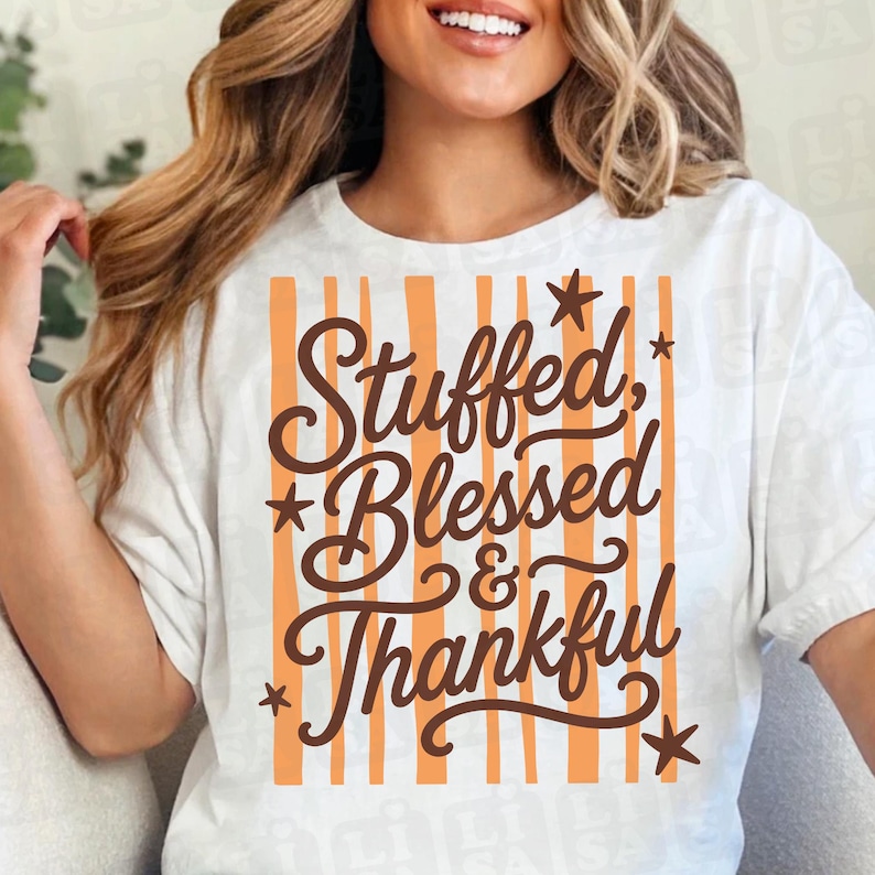 May include: A white short-sleeved t-shirt featuring a graphic design with vertical orange stripes. The text "Stuffed, Blessed & Thankful" is printed in dark brown cursive lettering over the stripes, accented by several small dark brown stars.