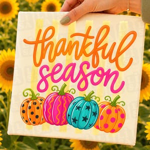 May include: A square canvas print featuring the text thankful season in orange and pink cursive script. Below the text are four pumpkins: an orange one with black dots, a pink one with orange stripes, a teal one with black stars, and an orange one with pink dots.
