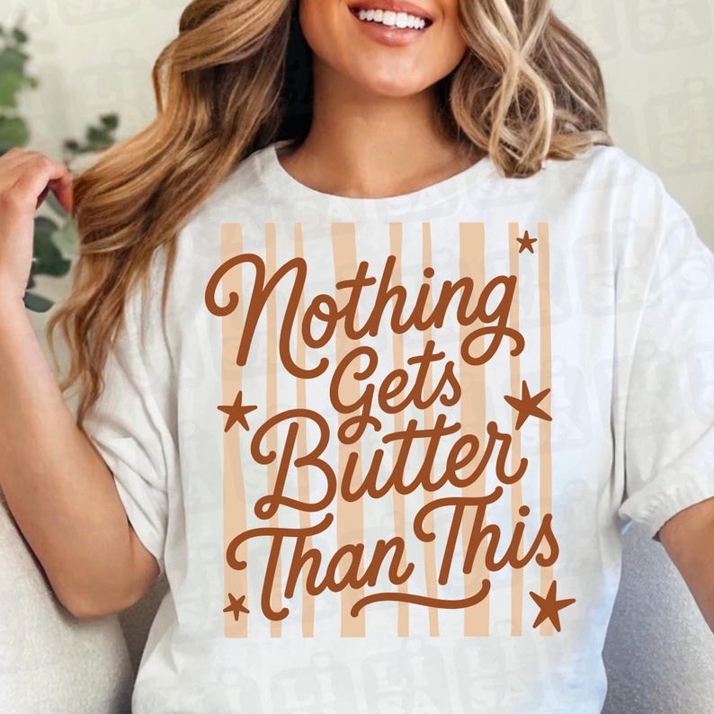 May include: A white short-sleeved t-shirt featuring a graphic design with vertical light tan stripes. The brown text reads Nothing Gets Butter Than This, accompanied by four small brown star icons arranged around the central lettering.